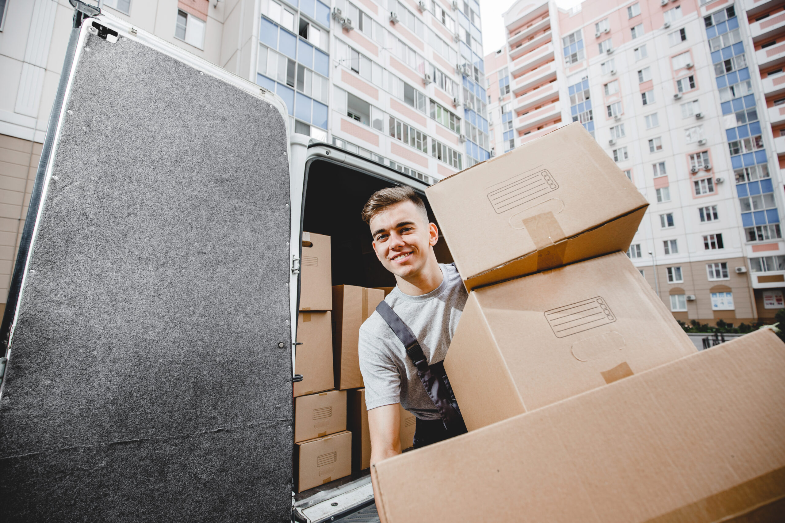 A young handsome smiling worker wearing uniform is unloading the van full of boxes. The block of flats is in the background. House move, mover service.